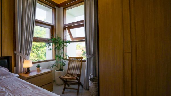 Cozy wooden bedroom corner with bed, nightstand lamp, potted plant, rattan chair, and large curtained windows overlooking trees.