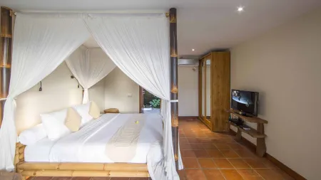 Cozy Balinese bedroom with bamboo four-poster bed, white canopy drapes, plush pillows, and wooden TV stand on terracotta tiles.