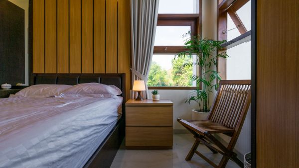 Cozy modern bedroom with wooden paneling, upholstered bed, bedside lamp, potted plant, folding chair, and large window with treetop view.