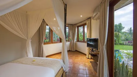 Elegant Balinese bedroom with white canopy bed, wooden floors, TV, and large windows overlooking tropical garden and infinity pool.