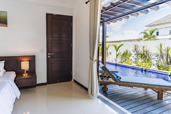 Luxurious tropical bedroom with king bed, bedside lamp, and open sliding doors revealing infinity pool and lounge chair.