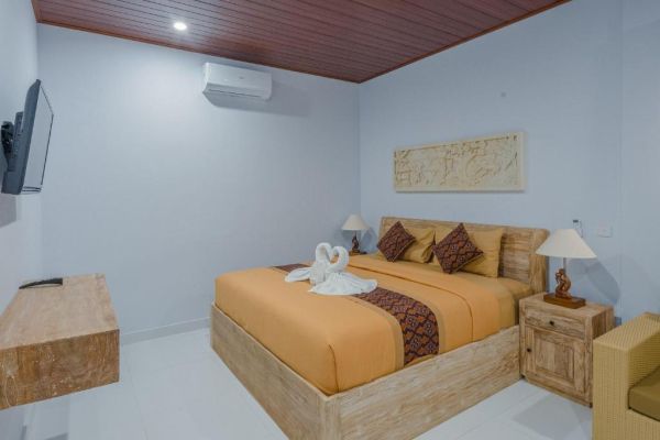 Cozy Balinese bedroom with wooden bed, orange batik bedding, swan towel, wall-mounted TV, and tropical decor.