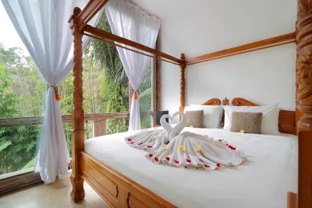 Luxurious tropical bedroom with carved wooden four-poster bed adorned by towel swan and frangipani flowers, overlooking lush green jungle through sheer-curtained windows. - Bali Villas