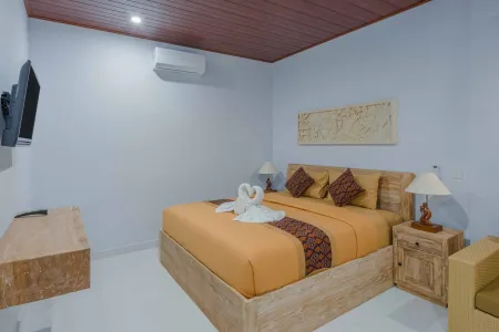 Cozy Balinese bedroom with wooden king bed featuring swan towel sculptures, orange batik bedding, mounted TV, and tropical wooden decor.