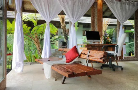 Rustic tropical workspace with wooden daybed, red pillow, iMac desk, white sheer curtains, and lush ferns in an open-air villa. - Bali Villas