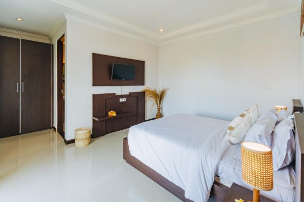 Spacious modern bedroom with white bedding, wooden platform bed, wall-mounted TV, rattan lamp, and tropical accents like frangipani flower and pampas grass in a Balinese-style villa.