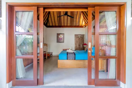 Spacious Balinese bedroom with open wooden sliding doors, thatched ceiling, blue king bed, towel swan, and tropical wooden decor.
