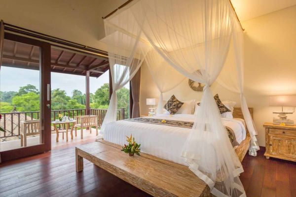 Luxurious Balinese villa bedroom with white canopy bed, wooden accents, and balcony view of tropical greenery.