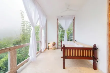 Tropical balcony bedroom with wooden daybed, sheer white curtains, ceiling fan, and misty jungle view.