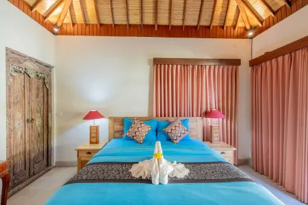 Cozy Balinese bedroom with turquoise bed, folded towel swan, carved wooden doors, orange curtains, and warm ambient lighting. - Bali Villas