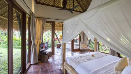 Luxurious tropical bedroom in a Bali villa with canopy bed, thatched roof, wooden accents, and garden views through open doors.