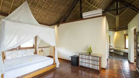 Luxurious tropical bedroom with white canopy bed, mosquito netting, thatched ceiling, wooden dresser, and open-plan bathroom in a Bali villa.