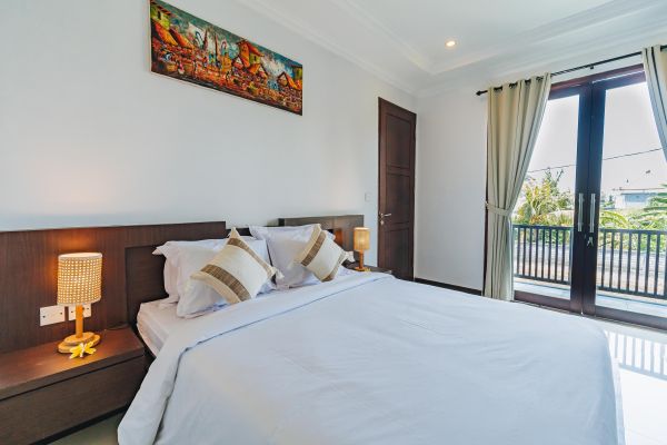 Spacious tropical bedroom with white king bed, rattan lamps, abstract wall art, and open sliding doors to palm-view balcony.