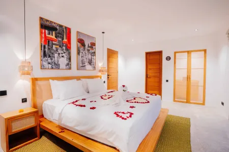 Romantic Balinese bedroom suite with wooden platform bed scattered in red rose petals, white linens, framed tropical artwork, and warm pendant lighting.