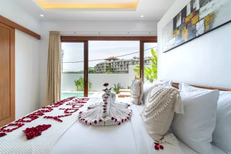 Romantic luxury bedroom in tropical villa with heart-shaped rose petals, towel swan on bed, bohemian pillows, and infinity pool view through sliding doors.