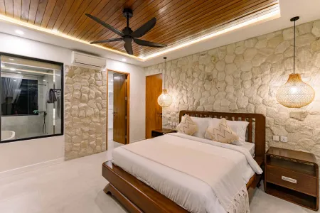 Cozy Balinese bedroom interior with wooden ceiling fan, textured stone walls, king bed in white linens, rattan pendant lights, and night view through window.