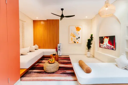 Modern tropical living room interior with white L-shaped sofa, wooden paneling, abstract floral art, potted plant, fruit basket on red rug, and wall-mounted TV showing fireplace. - Bali Villas