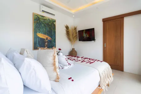 Romantic Bali bedroom with white bed scattered in red rose petals spelling "LOVE", pampas grass vase, surf painting, and wall-mounted TV.