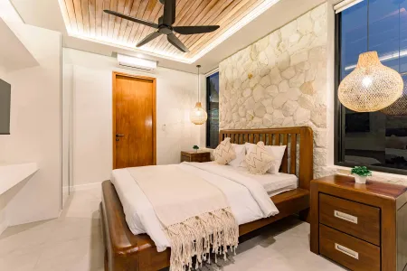 Cozy Balinese-style bedroom with wooden king bed, textured stone wall, woven pendant lights, and large window at dusk.