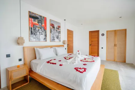 Romantic Balinese villa bedroom with heart-shaped red rose petals and towel swan on a white wooden bed, framed temple artwork, and soft tropical lighting.