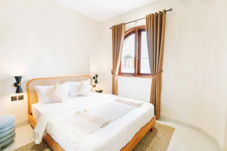 Cozy modern bedroom with wooden bed, white linens, arched window with brown curtains, and black wall sconces on light walls.
