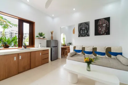 Modern minimalist open-plan kitchen and living area with wooden cabinets, Buddha wall art, beige daybed, and tropical plants. - Bali Villas