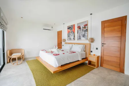 Luxurious tropical bedroom with platform bed adorned in rose petals and towel swan, abstract wall art, rattan chair, and ocean-view doors.