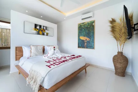 Romantic Bali honeymoon bedroom with wooden bed scattered in red rose petals spelling "HONEYMOON," tropical surf painting, abstract art, pampas grass, and wall-mounted TV.
