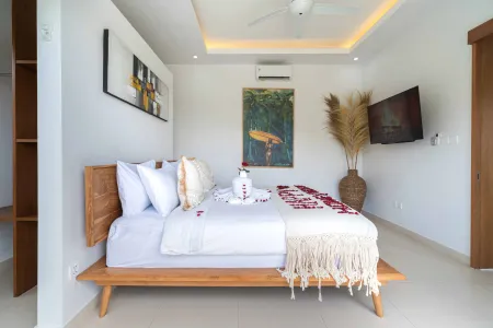 Luxurious tropical bedroom in a Bali villa featuring a wooden mid-century bed with white linens, scattered rose petals, wall art, pampas grass, and ceiling fan.