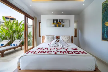 Romantic Bali honeymoon suite bedroom with a king bed adorned in red rose petals spelling "Happy Honeymoon," towel swans, and an open balcony overlooking tropical greenery.