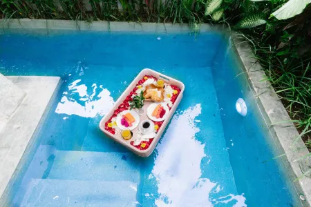 Wooden tray with tropical fruits, flowers, and plates floating in a clear blue infinity pool amid lush greenery. - Bali Villas