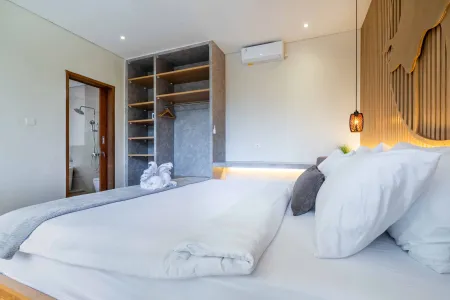 Modern tropical bedroom in a Balinese villa featuring a white king bed, open wooden wardrobe with towels, pendant lighting, and an adjacent shower bathroom.