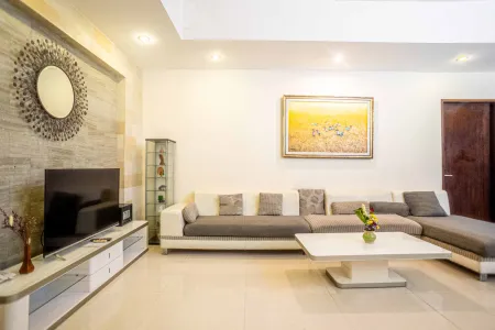 Modern living room interior with L-shaped gray sofa, wall-mounted TV, white coffee table, and decorative sunburst mirror on textured wall. - Bali Villas
