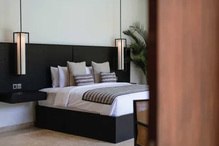 Modern tropical bedroom featuring a black headboard bed with white linens, pendant lights, potted palm, and wooden door.