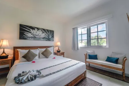 Cozy Balinese bedroom with wooden king bed, white linens, teal pillows, lotus wall art, and tropical window views.