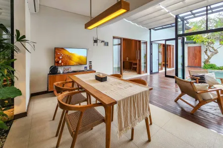 Modern tropical villa dining room with wooden table, chairs, wall-mounted TV, and open glass doors to wooden deck and infinity pool. - Bali Villas