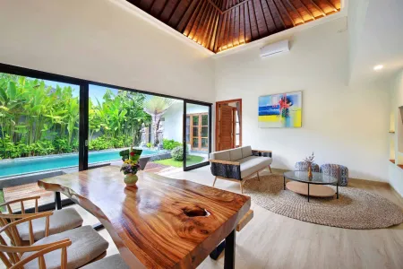 Modern Balinese villa interior featuring a natural wood dining table, lounge seating, abstract wall art, and panoramic views of lush tropical garden and infinity pool through floor-to-ceiling glass doors. - Bali Villas