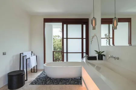 Luxurious modern bathroom with freestanding white bathtub, tropical balcony view, orchids, and wooden sliding doors. - Bali Villas