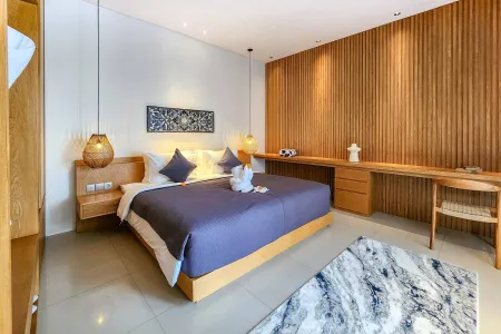 Modern Balinese bedroom interior with king bed in blue linens, swan towel, wooden panel walls, desk, and pendant lamps.