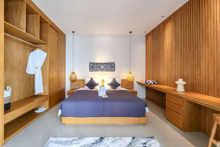 Spacious Balinese-style luxury bedroom with king bed in purple linens, towel swan, wooden wardrobe with robe, bamboo lamps, and built-in desk. - Bali Villas