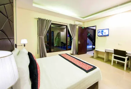 Spacious modern hotel bedroom with king-size bed, balcony overlooking tropical garden at night, desk, and wall TV.