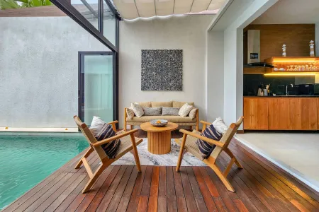 Modern poolside lounge in a tropical villa featuring beige sofa, wooden armchairs, and low table on a wooden deck, with glass walls and kitchen backdrop. - Bali Villas