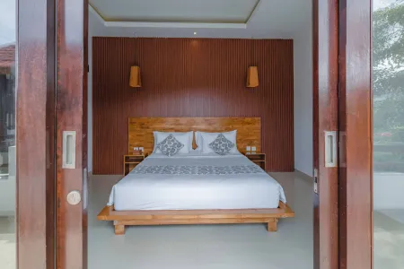 Spacious Balinese bedroom with wooden low-profile bed, white linens, open sliding doors to lush garden view, and warm pendant lighting.
