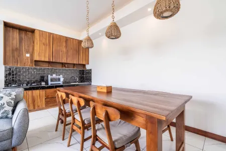 Modern Balinese-style kitchen with wooden cabinets, black tile backsplash, woven pendant lights, and teak dining table. - Bali Villas