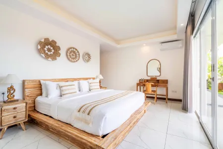 Spacious Bali-style bedroom with teak wood bed, white linens, woven wall decor, vanity desk, and sliding doors to tropical balcony.