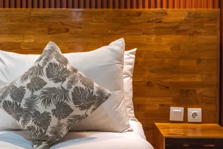 Cozy bedroom interior with slatted wooden headboard, white pillows in leaf pattern, and bedside lamp on wood nightstand.