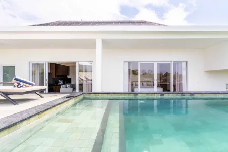 Modern white tropical villa with turquoise infinity pool, lounge chair, and open sliding doors revealing interior bedroom.