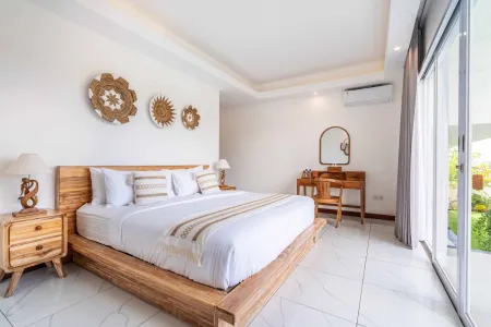 Bright, airy tropical bedroom with wooden platform bed, white linens, rattan wall art, vanity desk, and sliding doors to lush garden view.