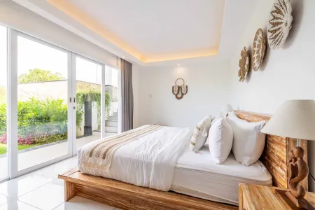 Spacious tropical bedroom with wooden platform bed, white linens, carved lamp on nightstand, and sliding doors opening to lush garden.
