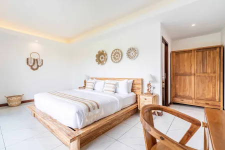 Spacious Balinese-style bedroom with wooden platform bed, white linens, woven wall art, and natural teak furniture.